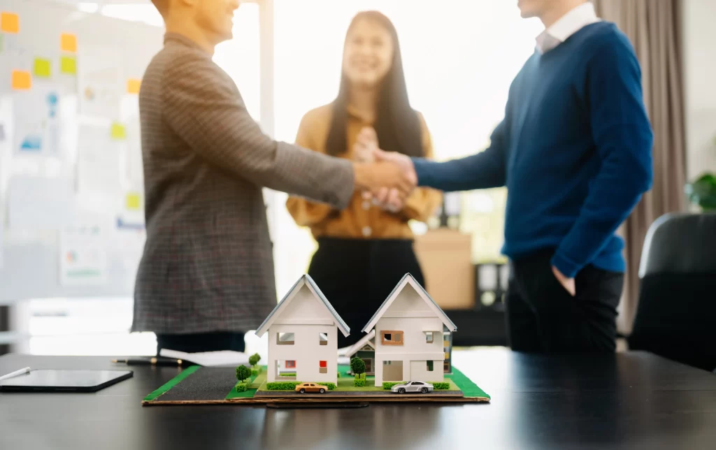 Dos personas estrechándose la mano frente a una mujer y una maqueta de casas,haciendo referencia inversión en franquicias en México