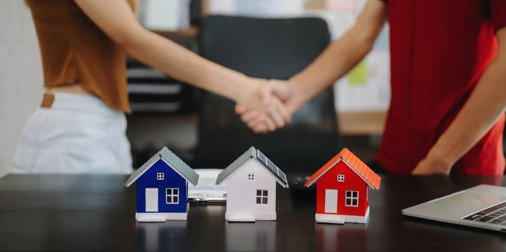 Manos estrechándose con casas miniatura. Simboliza acuerdo en franquicias inmobiliarias nacionales vs. internacionales: análisis para inversionistas.
