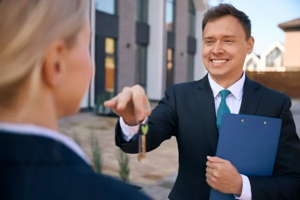 Un agente inmobiliario saluda cordialmente a una clienta frente a un edificio moderno, mientras le explica cómo funciona la inversión en bienes raíces y sus beneficios.