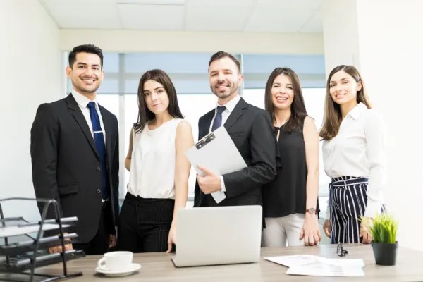 Cuatro personas vestidas de manera formal posan sonrientes frente a un escritorio en una oficina, mostrando un equipo unido que participa en los mejores negocios en México.