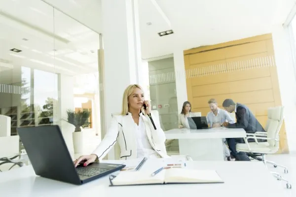 Una mujer habla por teléfono frente a una computadora en un entorno empresarial moderno, posiblemente atendiendo temas relacionados con cómo se administra una franquicia a distancia.