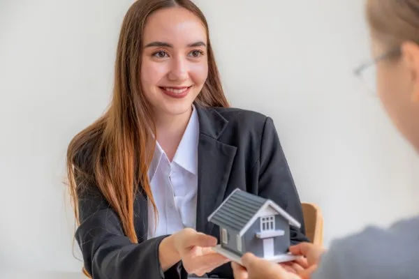 Una mujer con traje de oficina sostiene una pequeña figura de casa y documentos, lo que sugiere una presentación relacionada con qué se necesita para tener una franquicia en el sector inmobiliario.