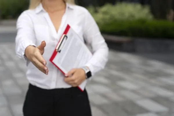 Una persona con camisa blanca ofrece un apretón de manos mientras sostiene una carpeta roja, gesto típico al cerrar un negocio seguro.